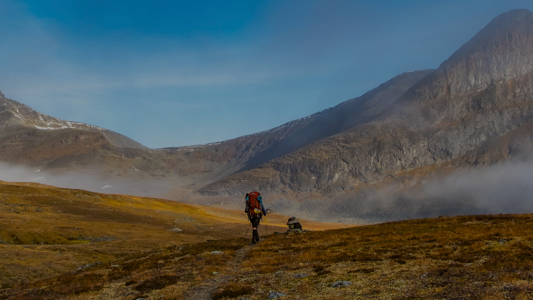 Wanderer mit großem Rucksack und Trekkingstöcken folgt schmalem Pfad durch herbstlich gefärbte Tundra im Sarek-Nationalpark, im Hintergrund steile, felsige Berge mit Restschnee und leichter Nebel unter blauem Himmel
