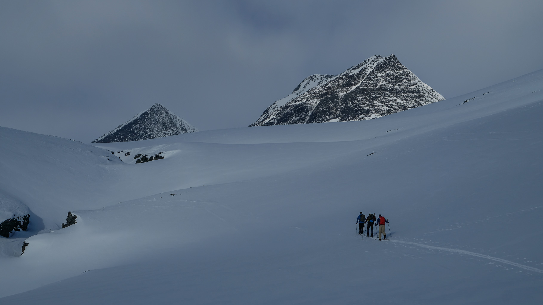Drei Skitrekkende steigen mit Pulka-Schlitten über weite, verschneite Hochebene im Fjell, im Hintergrund zwei markante, schneebedeckte Gipfel unter grauem Himmel.