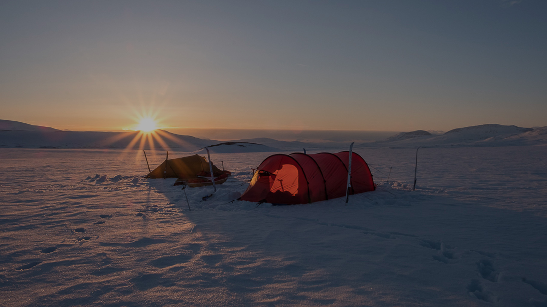 Zwei Zelte stehen im Schnee unter einer tiefstehenden Sonne, umgeben von weiten Hügeln