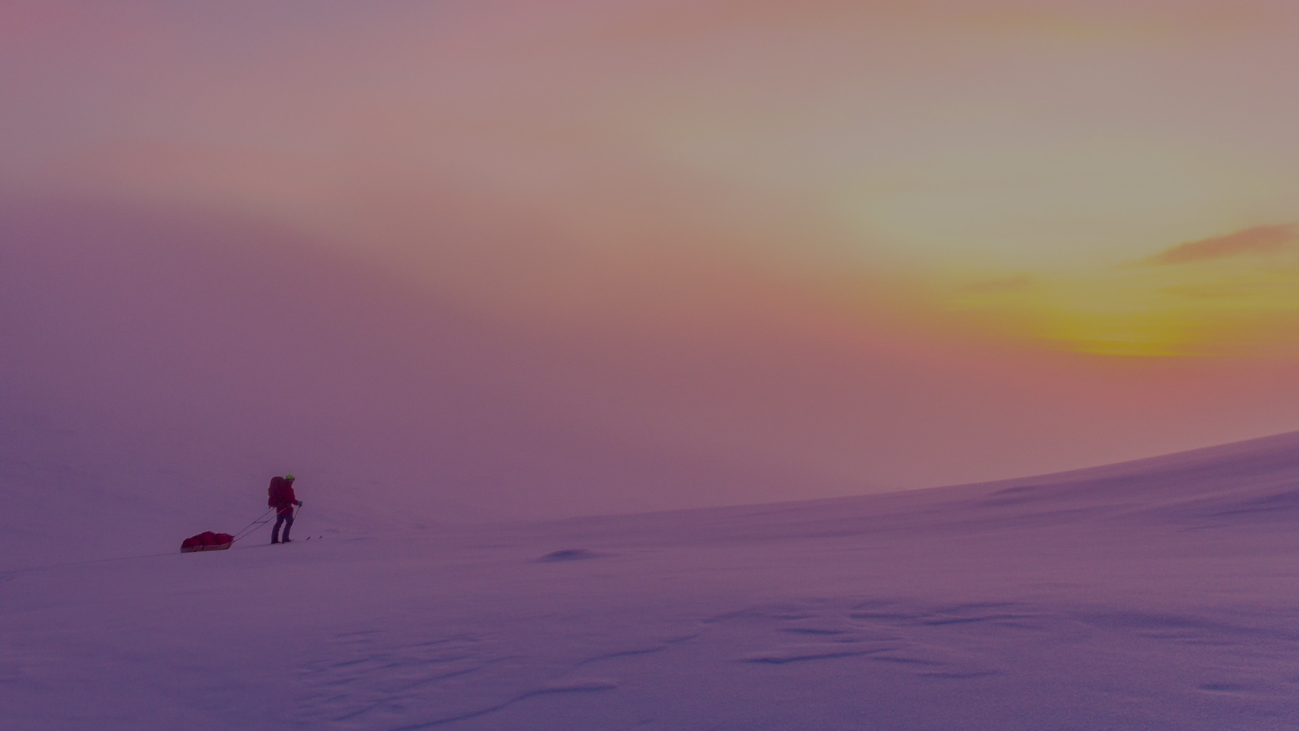 Person mit großem Rucksack zieht Pulka-Schlitten über weite, verschneite Hochebene im Fjell bei Sonnenuntergang; Himmel leuchtet in warmen Rosa-, Orange- und Gelbtönen, Schnee reflektiert das Licht sanft.