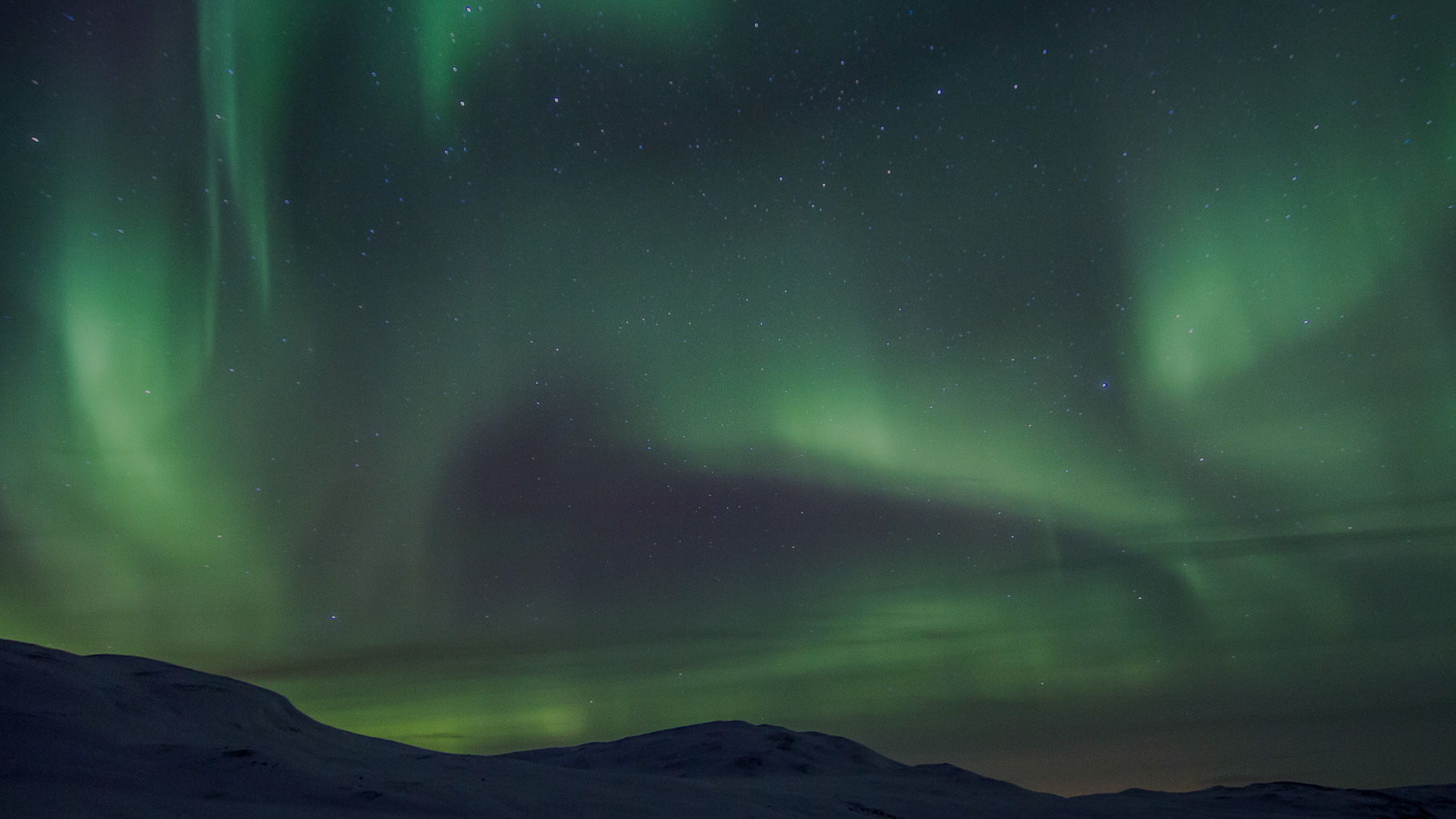 Grün leuchtende Nordlichter tanzen über dunklem Himmel mit vielen Sternen, darunter verschneite Hügel einer arktischen Landschaft im sanften Mondlicht.