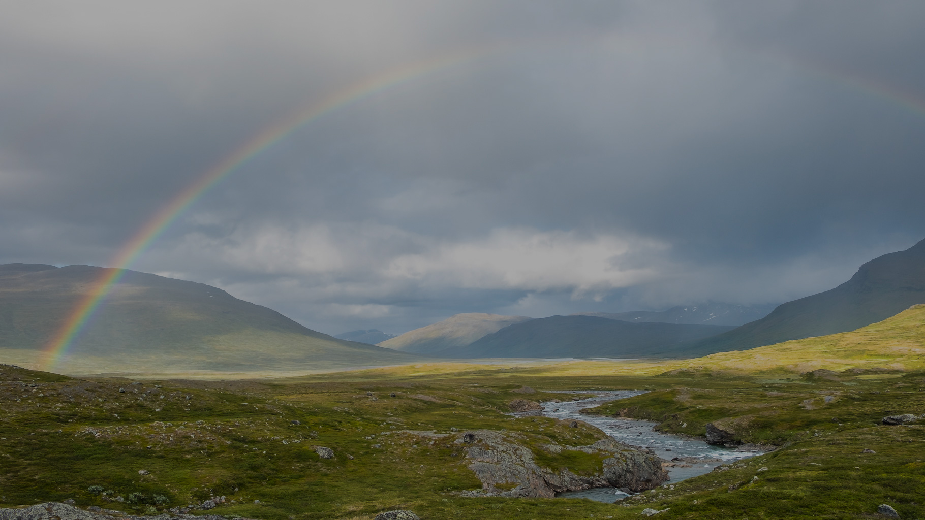 Weite grüne Tundralandschaft im Sarek-Nationalpark mit sanften Hügeln und einem klaren Fluss, darüber spannt sich ein Regenbogen unter dunklen Wolken, im Hintergrund leuchtet ein Sonnenfleck auf den Bergen.