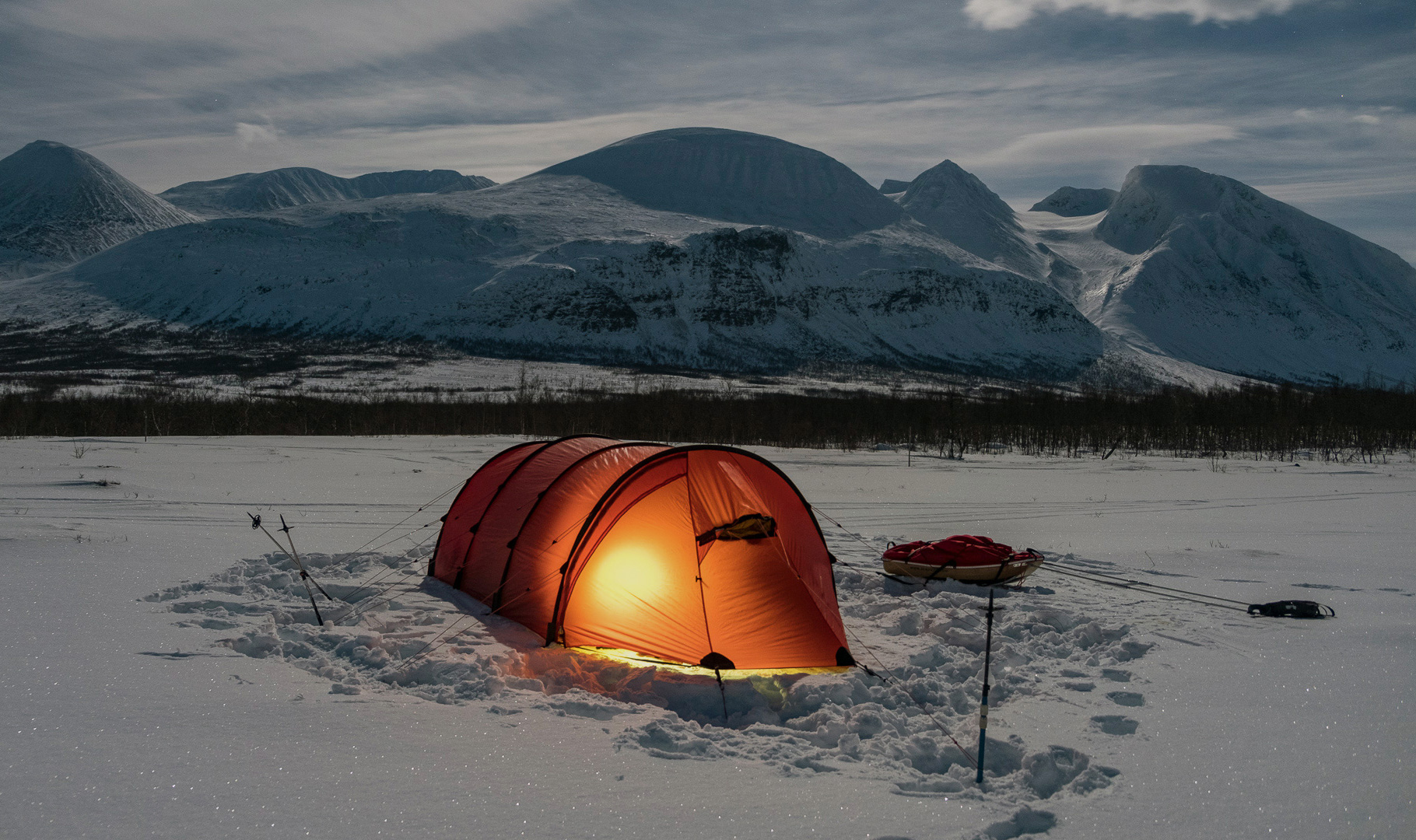 Leuchtend orangefarbenes Tunnelzelt steht im tiefen Schnee vor einer imposanten Bergkulisse in Lappland; das warme Licht aus dem Inneren kontrastiert mit den blauen Schatten der verschneiten Gipfel unter wolkigem Himmel.