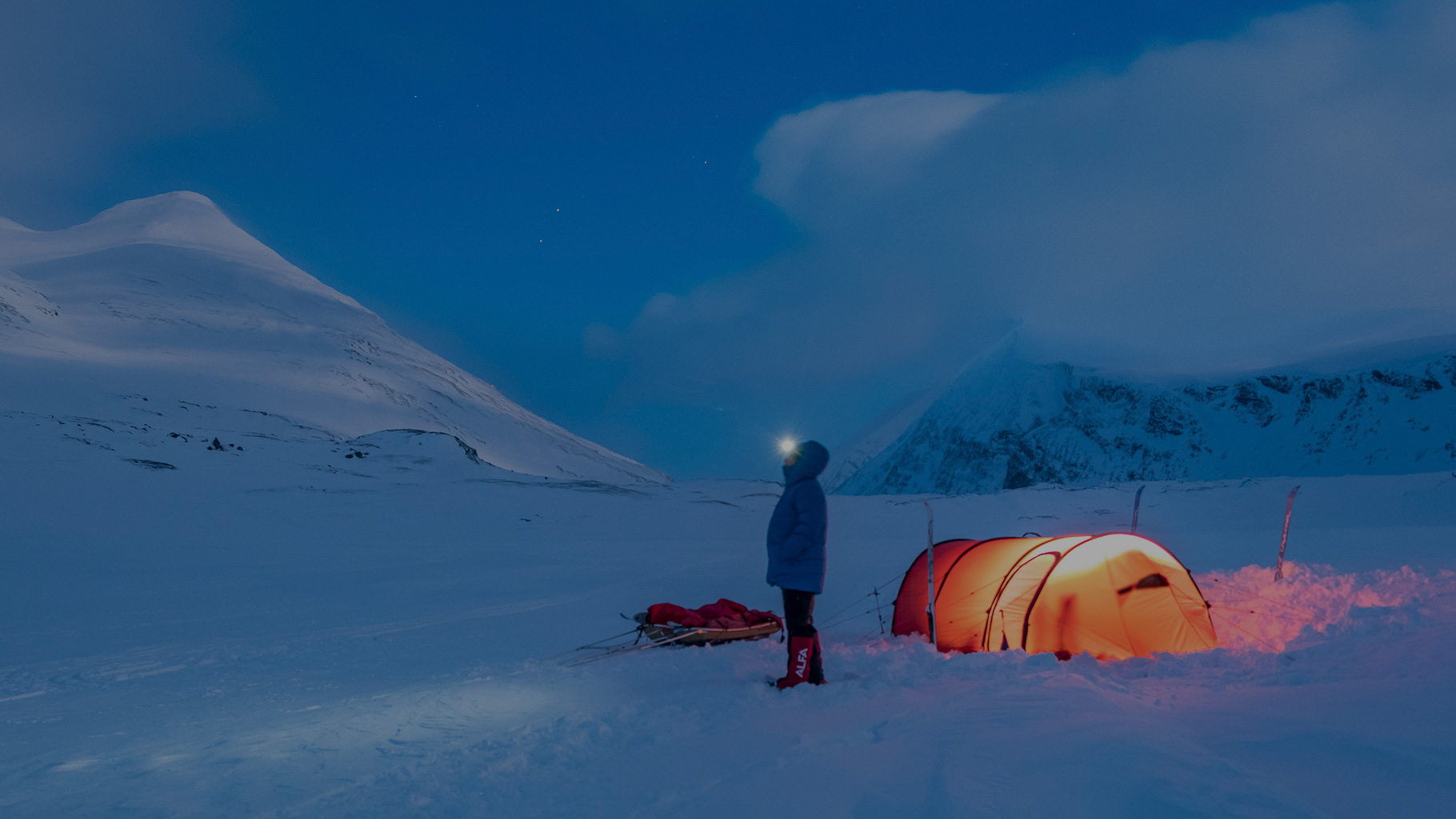 Person steht mit Stirnlampe neben orange leuchtendem Zelt im tiefen Schnee, umgeben von hohen, verschneiten Bergen unter dunklem, blauen Abendhimmel in Lappland.