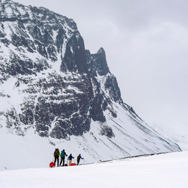 Eine kleine Gruppe von Skitrekking-Teilnehmern zieht Pulka-Schlitten über eine weite Schneefläche vor steiler Felswand im skandinavischen Gebirge.