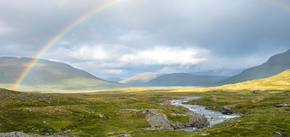 Ein Regenbogen spannt sich über ein grünes Tal mit Flusslauf zwischen sanften Bergen in Skandinavien, unter wechselhaftem Himmel mit Sonne und Wolken.