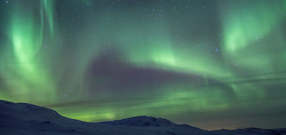 Grün-violette Polarlichter leuchten über verschneiten Hügeln in der skandinavischen Nacht, klarer Sternenhimmel im Hintergrund.