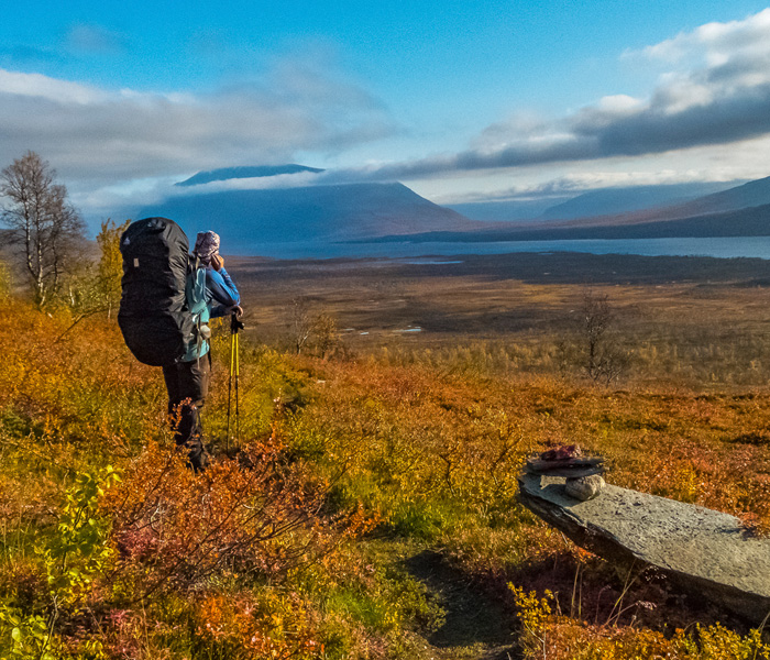 Wanderer mit großem Rucksack und Trekkingstöcken geht durch herbstlich gefärbte Fjelllandschaft im Sarek-Nationalpark, im Hintergrund weite Ebene mit See und Bergen unter blauem Himmel mit Wolken.