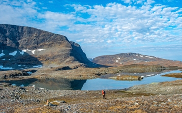 Weite Fjelllandschaft im Sarek-Nationalpark mit klaren Bergseen, Felsen und Schneeresten, im Vordergrund ein Wanderer mit rotem Rucksack unter blauem Himmel mit Schäfchenwolken.