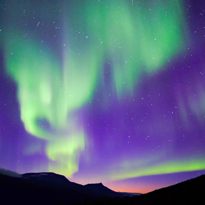 Grün-violette Polarlichter tanzen über dunklen Bergsilhouetten im Sarek-Nationalpark, klarer Sternenhimmel und zarter Dämmerung am Horizont.