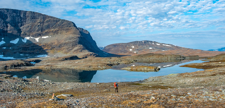 Wanderer mit rotem Rucksack steht am Ufer eines klaren Bergsees im Sarek-Nationalpark, umgeben von felsiger Tundra und schneebedeckten Gipfeln unter blauem Himmel.