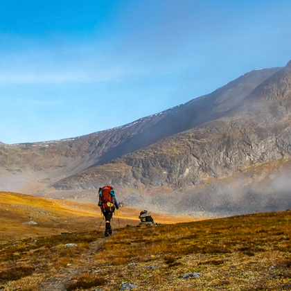 Wanderer mit rotem Rucksack geht über herbstlich gefärbte Fjelllandschaft im Sarek-Nationalpark, Nebelschwaden steigen vor steilen Berghängen auf unter klarem blauem Himmel.