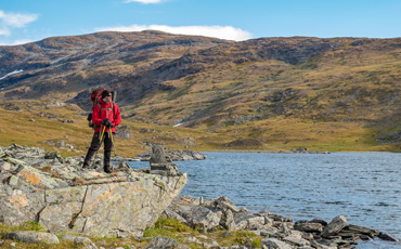 Wanderer mit rotem Rucksack steht auf Felsen am Ufer eines klaren Bergsees im Sarek-Nationalpark, umgeben von herbstlich gefärbten Hügeln und schneefreien Gipfeln unter blauem Himmel.