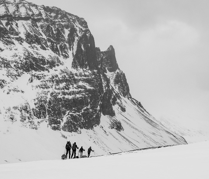 Vier Skitrekkende ziehen mit Pulka-Schlitten über weite verschneite Ebene unter steiler Felswand im Hochgebirge, grauer Himmel und dramatische Bergkulisse im Hintergrund.