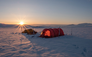 Zwei Zelte stehen im Schnee unter einer tiefstehenden Sonne, umgeben von weiten Hügeln