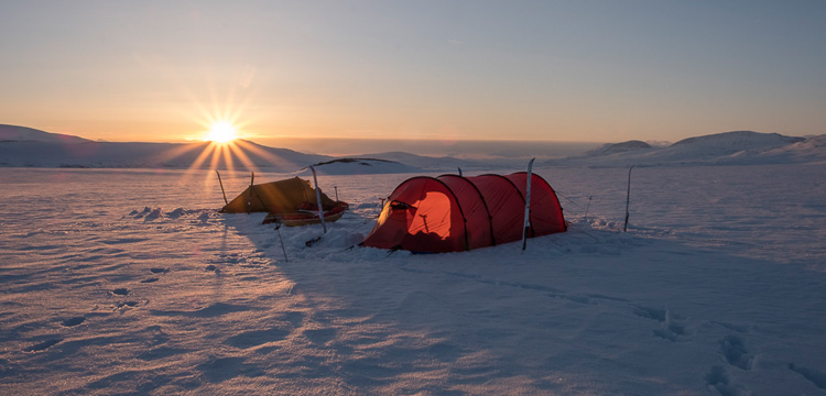 Zwei Zelte stehen im Schnee unter einer tiefstehenden Sonne, umgeben von weiten Hügeln
