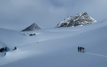 Vier Skitrekkende ziehen mit Pulka-Schlitten durch weite, verschneite Hochgebirgslandschaft im Narvikfjell, im Hintergrund ragen die Gipfel des Kebnekaise unter leicht bewölktem Himmel auf.