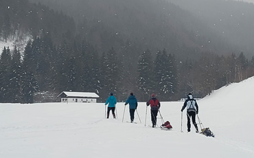 Vier Skitrekkende ziehen mit Pulka-Schlitten über verschneite Wiese bei Lenggries, im Hintergrund dichter Tannenwald und eine kleine Hütte unter grauem Winterhimmel.