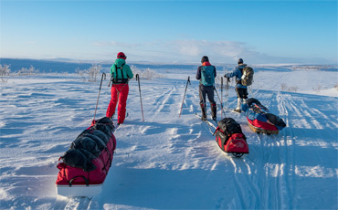 Vier Skitrekkende ziehen mit Pulka-Schlitten über weite, verschneite Ebene im Fjell bei Lenggries, im Hintergrund sanfte Hügel unter blauem Himmel mit klarer Sicht.