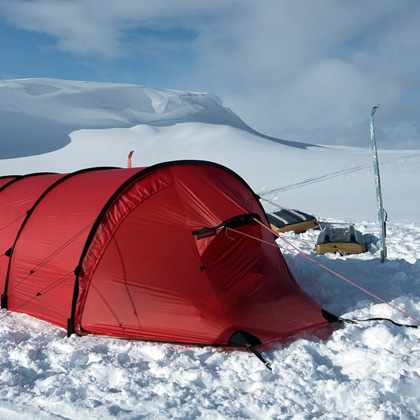 Rotes Tunnelzelt steht im tiefen Schnee auf einer Hochebene, daneben Ausrüstung und Skistöcke, im Hintergrund verschneite Hügel unter blauem Himmel mit leichten Wolken.