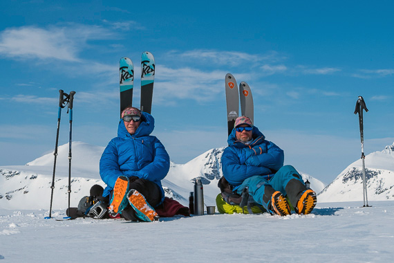 Corry Heiland und Mark Renker von Wildtreks sitzen in blauen Daunenjacken im Schnee mit Tourenski und Stöcken, umgeben von verschneiten Bergen unter blauem Himmel.