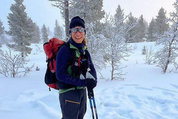 Eine Frau, Corry Heiland von Wildtreks steht mit rotem Rucksack und Skistöcken lächelnd im Fjell, umgeben von schneebedeckten Bäumen unter blassem Winterhimmel.