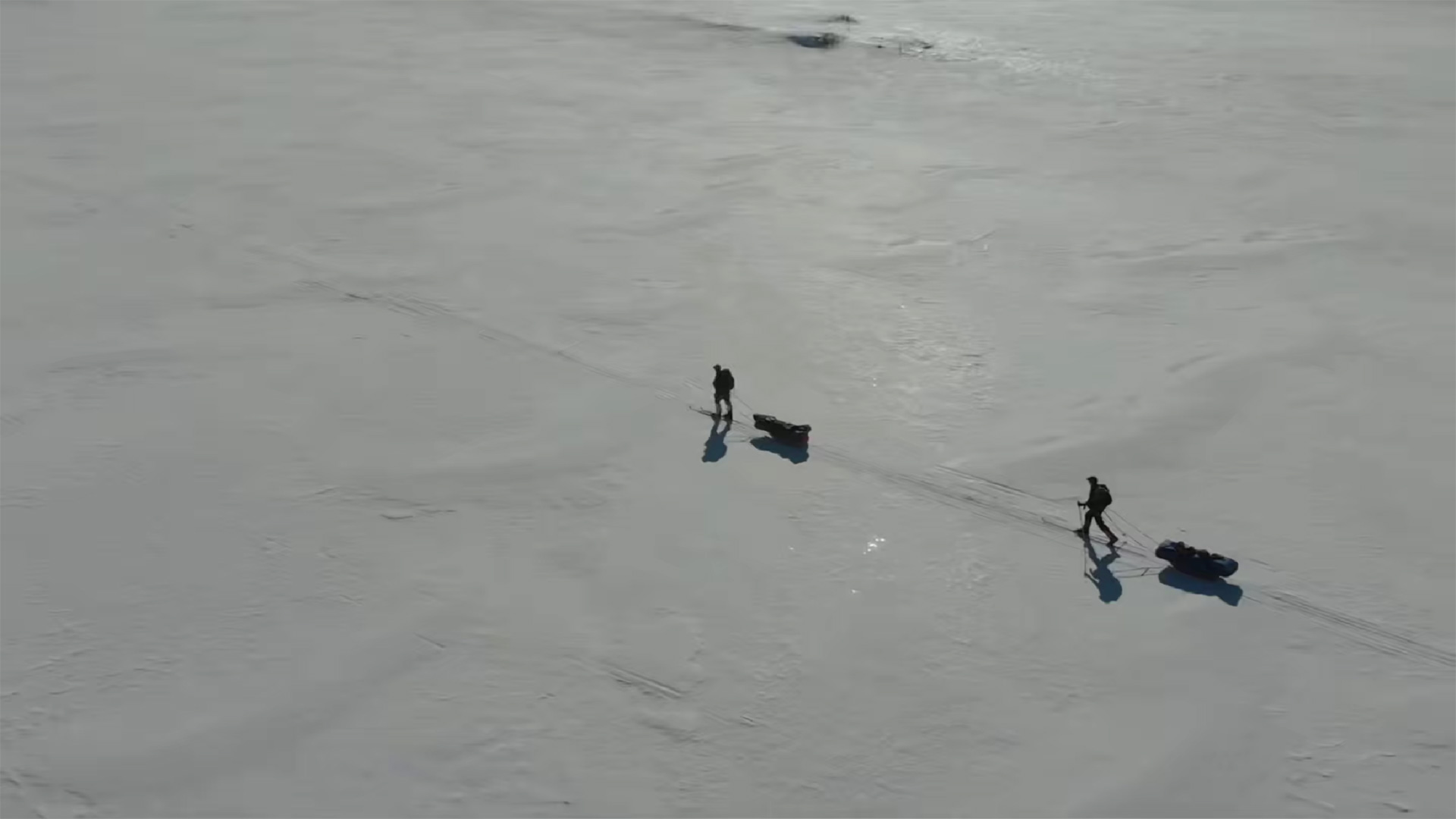 Drohnenaufnahme von zwei Skitrekkenden, die mit Pulka-Schlitten über eine weite, verschneite Ebene ziehen; lange Schatten fallen über die glatte Schneefläche unter klarem, blauem Himmel.
