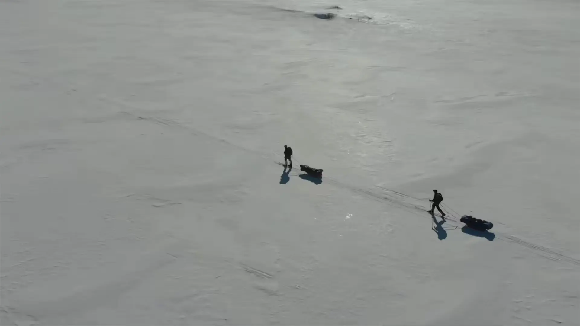 Drohnenaufnahme von zwei Skitrekkenden, die mit Pulka-Schlitten über eine weite, verschneite Ebene ziehen; lange Schatten fallen über die glatte Schneefläche unter klarem, blauem Himmel.
