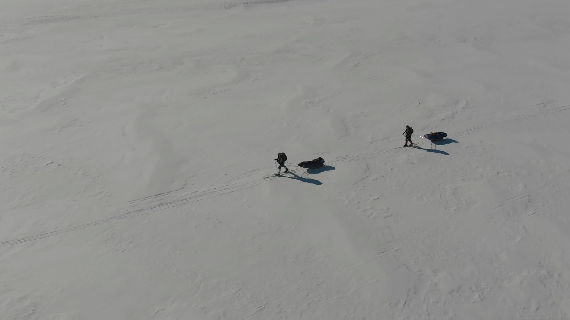 Drohnenaufnahme von zwei Skitrekkenden, die mit Pulka-Schlitten über eine weite, verschneite Ebene ziehen; lange Schatten fallen über die glatte Schneefläche unter klarem, blauem Himmel.