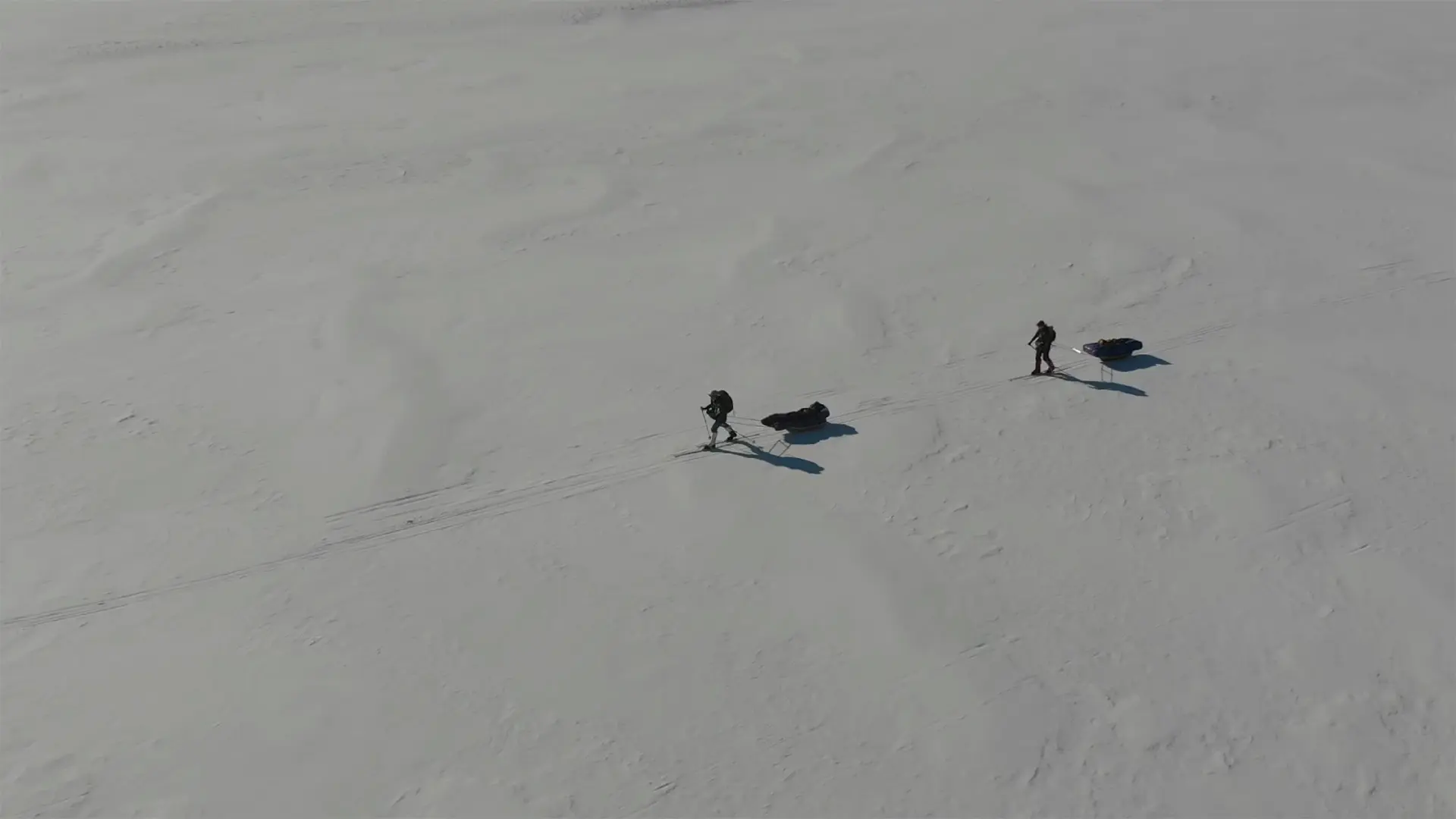 Drohnenaufnahme von zwei Skitrekkenden, die mit Pulka-Schlitten über eine weite, verschneite Ebene ziehen; lange Schatten fallen über die glatte Schneefläche unter klarem, blauem Himmel.